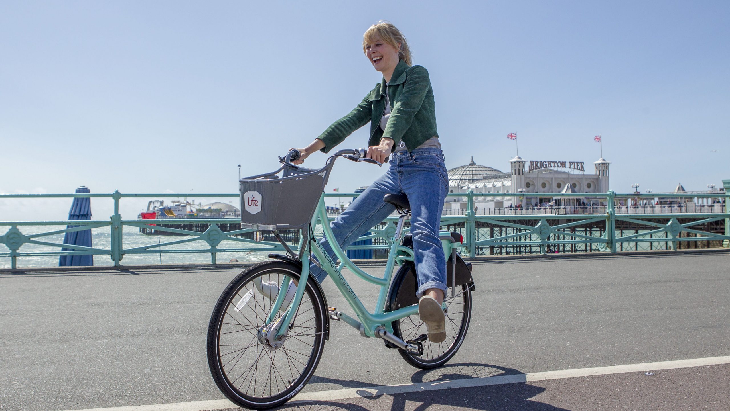 A picture of a woman riding a bike down Brighton Beach, with the pier in the background.